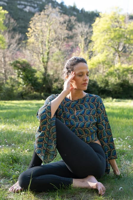 Myriam, fondatrice d'Essence de Toi, assise dans l'herbe en pratique de yoga, une main près du visage, les yeux fermés